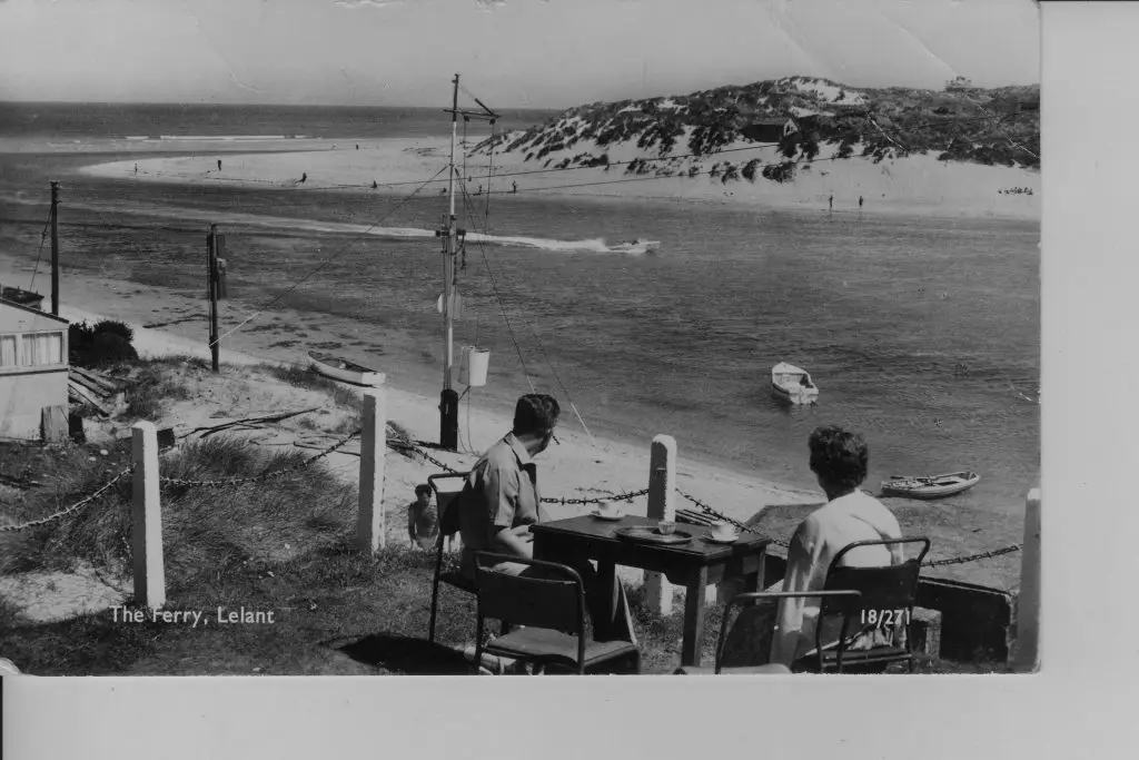 Black and White Photo of two people sitting at a table overlooking the beach with a small speed boat and two rowing boats in the water